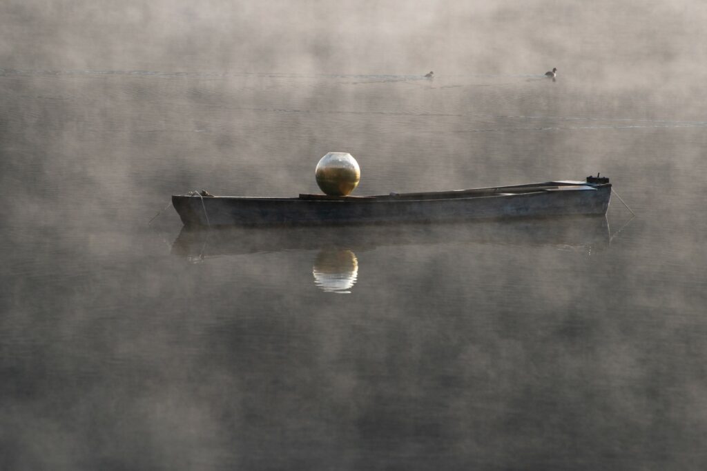 The wooden boat is on the foggy water. On board is a large shimmering glass bowl. A pair of water birds swim in the background.